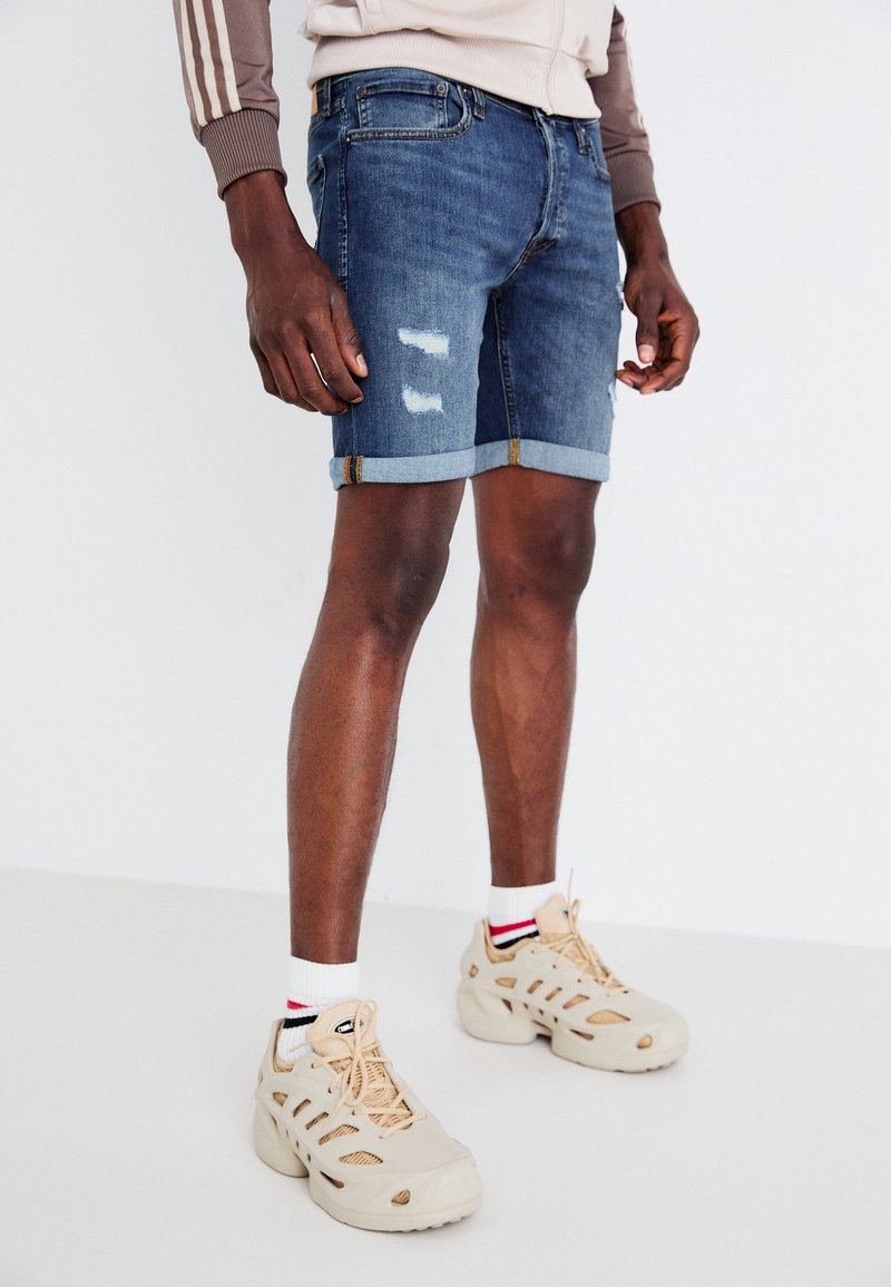 Jeansshorts in mittlerem Waschung mit abgetragenen Details und umgeschlagenen Säumen. Kombiniert mit beigen Sneakers und gestreiften Socken.