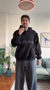 Černá fleecová mikina s předním černým panelem, opatřená kapucí na šňůrku a elastickými manžetami, spárovaná s tmavými džínami. Interiér.