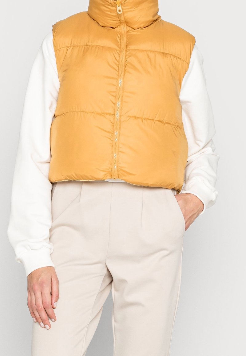 Femme portant une doudoune courte jaune moutarde par-dessus un top blanc à manches longues et un pantalon beige taille haute, une main dans la poche.