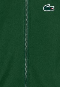 Zelena jakna na zip od teksturiranog materijala, sa središnjim rajsferšlusom i malim Lacoste logotipom u kontrastnim bojama s lijeve strane.