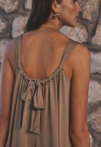 Robe longue et fluide en tissu taupe doux, présentant un dos froncé avec un détail en nœud, des bretelles larges et une texture lisse.