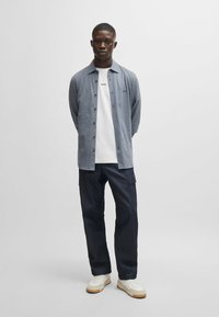 Camisa de botones gris sobre una camiseta blanca, combinada con pantalones cargo de mezclilla oscura y zapatillas blancas. El atuendo tiene un aspecto casual y en capas.