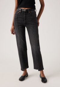 Kvinde iført sorte, højtaljede jeans med lige ben og sort bælte, kombineret med sorte loafers og en sort cropped top, stående.