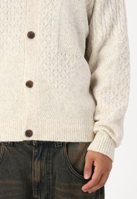 Beige stickad cardigan med ett strukturerat mönster, med träknappar och ribbade ärmslut; bärs över mörka byxor.