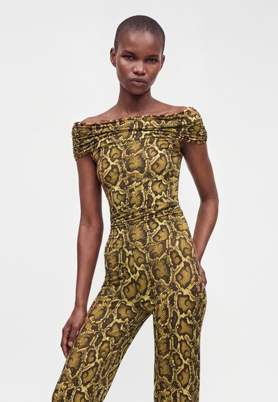 Femme mince aux cheveux courts portant une combinaison jaune et marron à imprimé serpent décolletée, debout devant un fond clair uni.