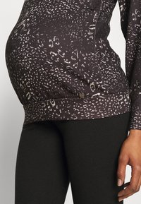 Zwangertop in donkerbruin met een subtiel wit abstract patroon, voorzien van een gathering aan de onderkant en een metalen accent aan de onderrand.