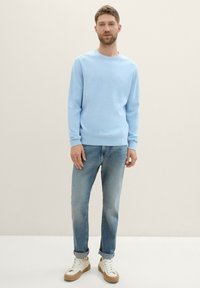 Hellblauer Pullover aus weichem Stoff, runder Ausschnitt und lange Ärmel, kombiniert mit faded blauen Jeans und weißen Sneakers mit braunen Sohlen.