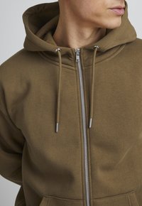 Brauner Reißverschluss-Hoodie mit einer vorderen Tasche, Zugbändern und silberfarbener Hardware. Hergestellt aus weichem Stoff mit glatter Textur. Das Design des Hoodies umfasst einen abgerundeten Halsausschnitt.
