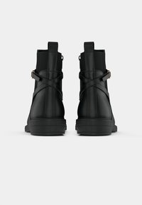 Par črnih gležnarjev s križnimi trakovi in pomočnimi zankami, prikazanih zadaj na enobarvni sivi ozadju.