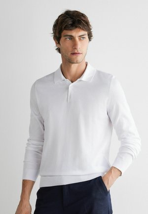 Mann mit kurzen braunen Haaren, der ein weißes langärmliges Poloshirt und dunkle Hosen trägt, schaut seitlich, mit einer Hand in der Tasche.