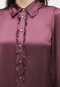 Μωβ σατέν μπλούζα με βολάν και μικτούς ασημένιους και χρυσούς κουμπότρυπες, με μυτερό γιακά και μακριά μανίκια.