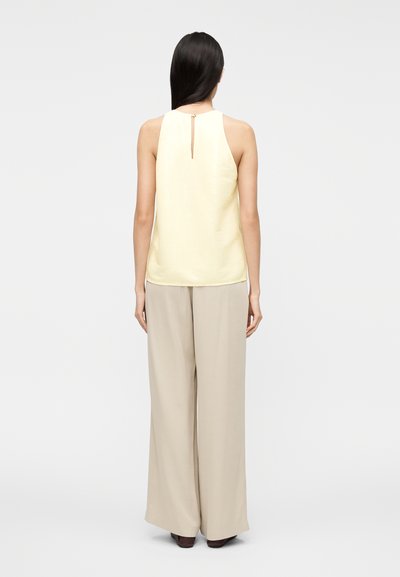 Κίτρινο τοπ χωρίς μανίκια με λεπτομέρεια άνοιγμα στην πλάτη, συνδυασμένο με μπεζ παντελόνι φαρδιάς γραμμής, εμφανίζεται από πίσω σε απλό φόντο.