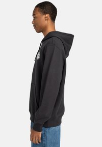 Black hoodie made of cotton, featuring a spacious kangaroo pocket, ribbed cuffs, and a logo print on the chest. Side profile view.
