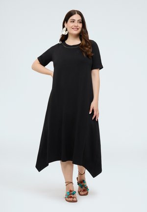 Vrouw met lang bruin haar draagt een zwarte midi-jurk met korte mouwen en veelkleurige pomponsandalen, staand met één hand op haar heup.
