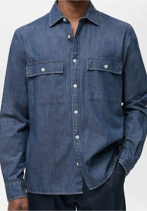 Homme portant une chemise en denim bleu à manches longues avec deux poches poitrine boutonnées et des boutons blancs, une main dans la poche.