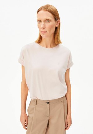 Woman with straight red hair wears a pale pink short-sleeve shirt tucked into beige high-waist trousers, standing against a white background.