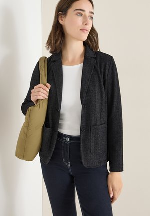 Blazer noir à chevrons avec revers, poches, porté sur un haut blanc, assorti à un jean foncé, tenant un sac à bandoulière vert olive.