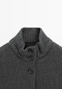Gilet en maille gris avec un col haut et côtelé, et trois boutons noirs bien visibles à l'avant. Texture douce et design sans couture.