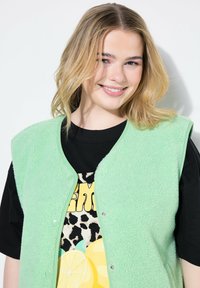 Mintgrön fleecest vest med knäppknappar, lagd över en svart t-shirt med grafiska tryck av gula citroner och leopardmönster.