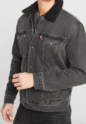 Mand iført sort denimjakke med sort sherpa-krave, knappet front, brystlommer og lange ærmer, stående mod en ensfarvet baggrund.