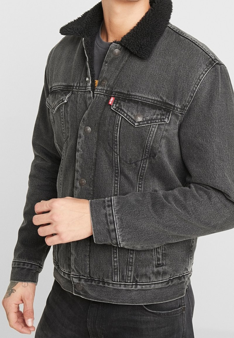 Homme portant une veste en denim noire avec col sherpa noir, devant boutonné, poches poitrine et manches longues, debout devant un fond uni.