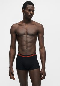 Άνδρας με μυώδη σωματότυπο που φοράει μαύρα μπόξερ με κόκκινη ζώνη μέσης «HUGO», στέκεται μπροστά σε απλό φωτεινό φόντο.