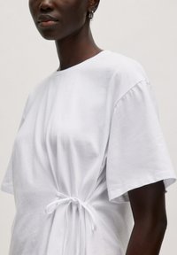 Witte katoenen t-shirt met korte mouwen, ronde halslijn en geplooide details bij de taille, vastgebonden met een witte strik. Glad textuur.