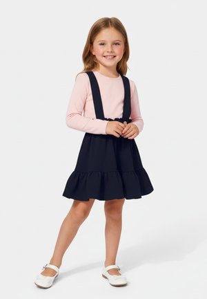 Granatowa sukienka z regulowanymi ramiączkami, falbanowanym dołem, zestawiona z jasnoróżową bluzką z długim rękawem oraz białymi butami z kokardkami.