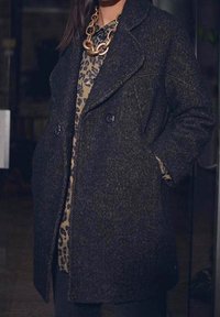 Cappotto doppiopetto in misto lana di colore grigio scuro con finitura testurizzata, dotato di rever a punta e tasche laterali, indossato sopra una maglia con stampa leopardata.