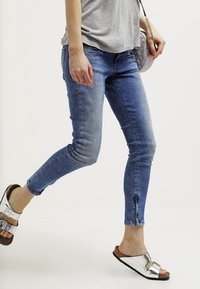 Skinny blue denim jeans with a faded finish, featuring a zip detail at the ankles. Paired with silver and brown slide sandals.