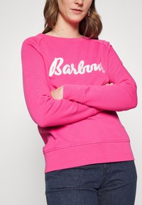 Sweat-shirt rose avec logo blanc "Barbour", col rond et poignets côtelés. Le tissu est doux, avec une texture lisse et une coupe décontractée.