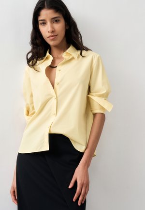Femme portant une chemise jaune pâle à boutons avec les manches retroussées et une jupe noire, debout devant un fond clair uni.