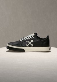 Μαύρο χαμηλό sneaker με άσπρα κορδόνια, άσπρη σόλα και λευκό λογότυπο με σταυρωτά βέλη στο πλάι, παρουσιάζεται σε ουδέτερη επιφάνεια.