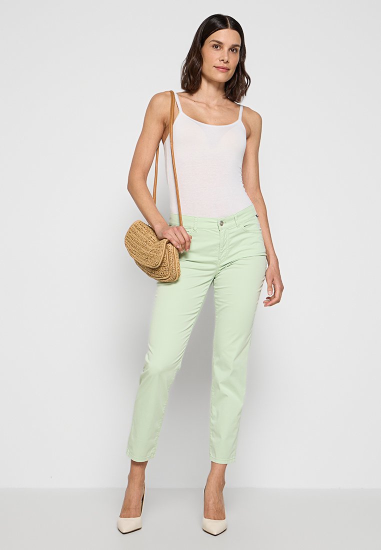 Hellgrüne Baumwollhose mit schmalem Schnitt, kombiniert mit einem weißen Tanktop und beigen High Heels. Eine gewebte braune Tasche wird schräg über die Schulter getragen.