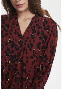 Bordeauxrote Bluse mit V-Ausschnitt, verziert mit einem schwarzen und beigen Leopardenmuster. Weicher, strukturierter Stoff mit langen Ärmeln und entspannter Passform.