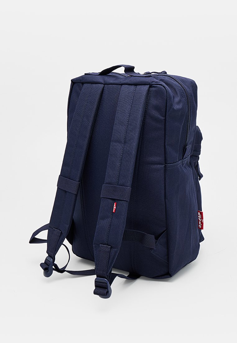 Mochila azul marino con correas acolchadas para los hombros, asa para llevar en la parte superior, bolsillo lateral y pequeña etiqueta roja de la marca en el lateral.
