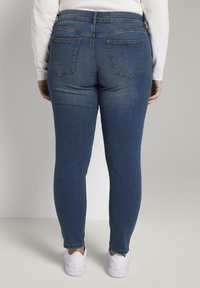 Jeans ajustados en denim azul medio con una textura suave, estilo de cinco bolsillos y detalle en el canesú trasero, combinados con zapatillas blancas.