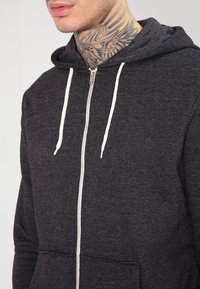 Grauer Zip-Hoodie aus weichem Stoff, mit einer Fronttasche, Kordelzügen und einer mittelhohen Kapuze. Auffälliges weißes Reißverschlussdetail.