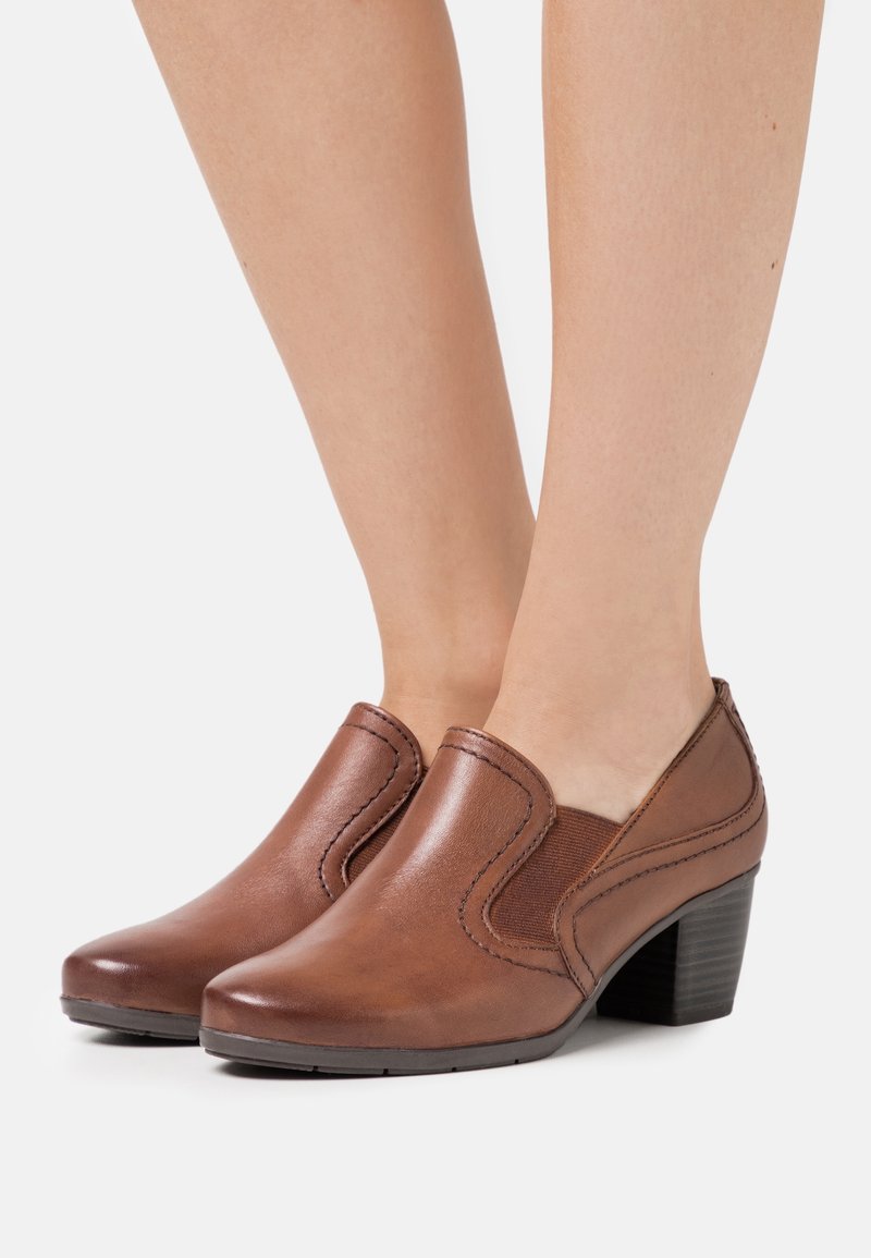 Chaussures à enfiler en cuir marron avec des talons empilés bas, portées pieds nus sur un fond blanc uni.