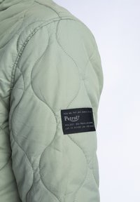 Ärmel einer hellgrünen, gesteppte Jacke mit einem strukturierten Wellenmuster, das ein schwarzes Markenetikett mit weißen Buchstaben auf dem Arm zeigt.