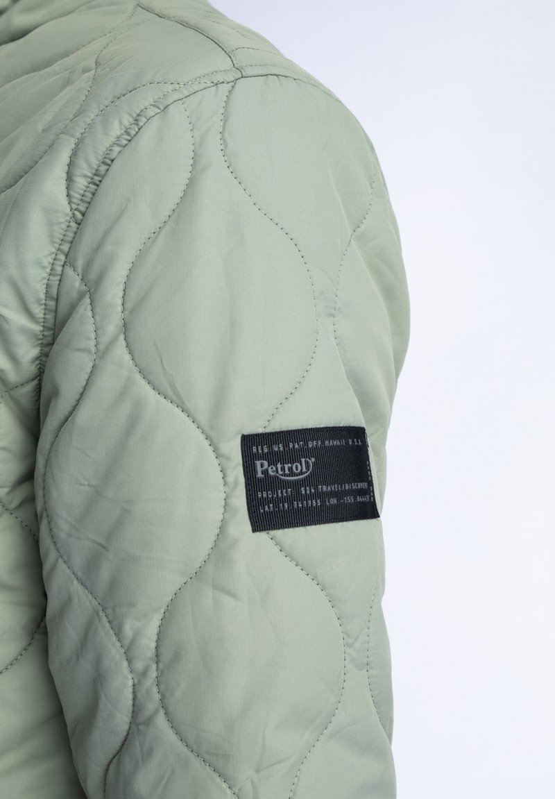 Manga de chaqueta acolchada verde claro con un patrón de olas texturizado, que presenta una etiqueta de marca negra con letras blancas en el brazo.