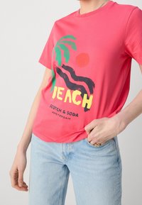Camiseta de algodón color rosa coral con un gráfico de una palmera negra y un sol, donde se lee la palabra "PLAYA" en amarillo. Corte relajado y escote redondo.
