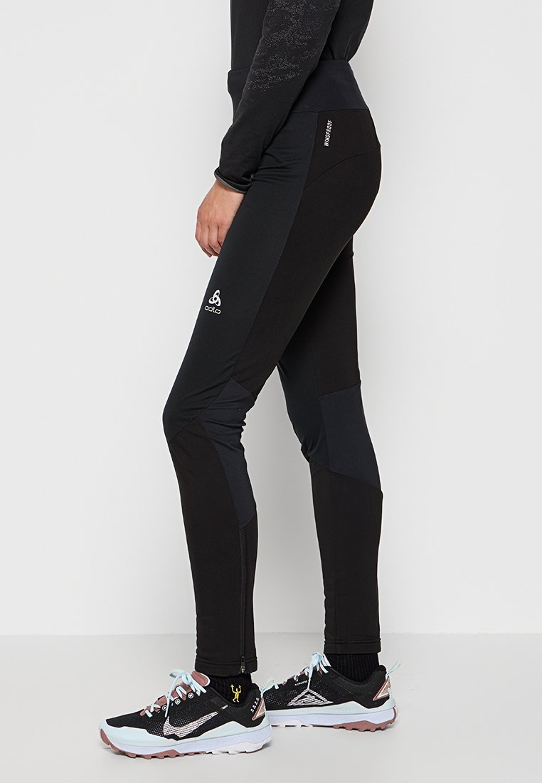 Nahaufnahme einer Person in schwarzen, winddichten Odlo-Sportleggings und schwarzen, weiß-rosa Trailrunningschuhen mit blauen Schnürsenkeln.