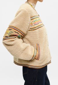 Beżowa puszysta kurtka z kolorowym haftem na klatce piersiowej i rękawach. Posiada zapięcie na guziki oraz kieszenie boczne. Teksturowany i przytulny fason.