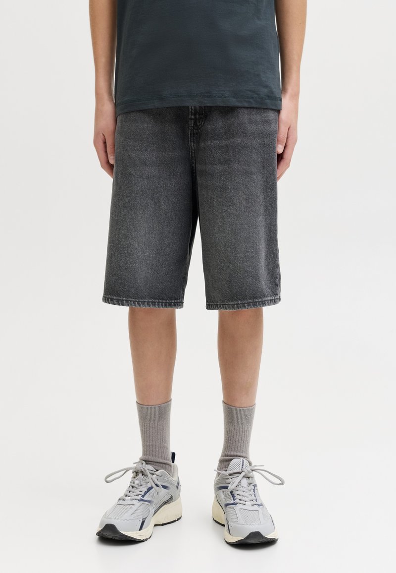 Persona che indossa pantaloncini di jeans neri al ginocchio, calzini grigi e scarpe sportive bianche, in piedi davanti a uno sfondo semplice.