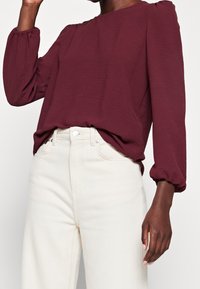 Blouse texturée bordeaux avec manches ballon, associée à un jean crème taille haute. La blouse a un col rond et une coupe décontractée.