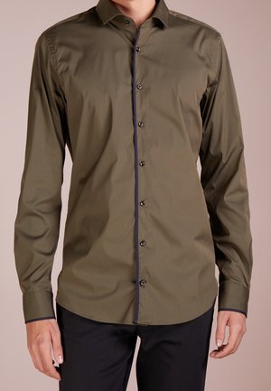 Homme portant une chemise boutonnée à manches longues vert olive avec une bordure sombre sur la ligne des boutons et les poignets, associée à un pantalon noir.