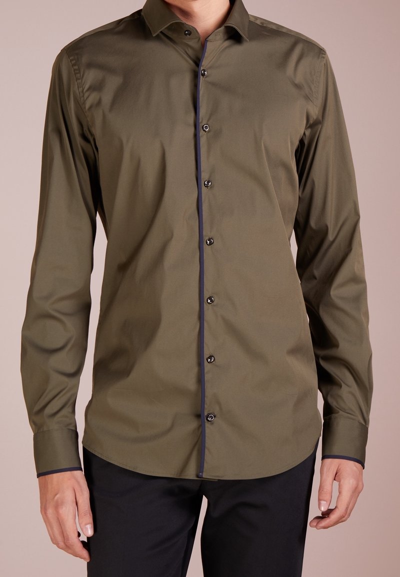 Homme portant une chemise boutonnée à manches longues vert olive avec une bordure sombre sur la ligne des boutons et les poignets, associée à un pantalon noir.