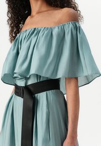 Abito off-the-shoulder in tessuto blu chiaro con un top volante arricciato, cintura nera cinchata in vita e dettagli di gonna plissettata.