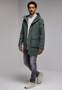 Green waterproof jacket with a hood, two front pockets, and snap buttons. Worn over a light gray sweater and paired with gray jeans and sneakers.